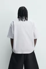 Camisa polo boxy fit de manga corta, confeccionada en tejido de algodón con diseño de rayas verticales celestes y blancas. Cuenta con cuello solapa, cierre frontal con botones y estampado frontal de texto en tono marrón con la leyenda "Poetic Work Since 1975".