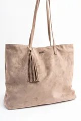 Bolso tipo tote de gran tamaño confeccionado en gamuza sintética color tostado. Presenta un diseño minimalista con asas largas de hombro y una pequeña placa metálica frontal con el logo de la marca.