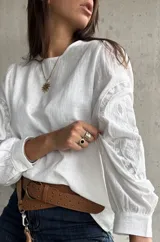 Blusa blanca de lino con mangas largas abullonadas, destacada por paneles laterales bordados con detalles de encaje. Presenta un corte holgado y cuello redondo.