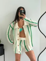 Conjunto de dos piezas tejido en crochet, compuesto por un cárdigan de manga larga con cuello tipo polo y cierre frontal de botones, y un short de tiro alto con cintura elástica. El diseño presenta un patrón de rayas verticales en color verde sobre una base color crema.