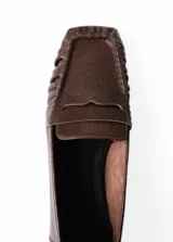 Mocasines de cuero color marrón oscuro, con diseño clásico de tira en el empeine, costuras fruncidas alrededor de la puntera cuadrada y suela plana.