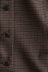 Chaleco largo sin mangas, con cuello redondo y cierre frontal de botones. Presenta un estampado de pata de gallo en tonos marrones y negros, bolsillos delanteros ribeteados y abertura en la parte trasera inferior.