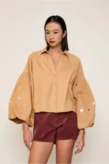 Camisa color beige de corte crop, con cuello camisero, mangas largas abullonadas con detalle de espejos y cierre frontal con botones.