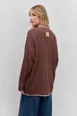 Cardigan de punto con diseño de trenzas, color marrón, con cierre frontal de botones y detalle de borde en color blanco. Presenta bolsillos frontales y mangas largas.