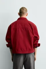 Cazadora de corte boxy fit en color bordó, confeccionada en tejido de algodón con efecto lavado. Presenta cuello solapa, cierre frontal con cremallera oculta y alamares metálicos decorativos. Incluye bolsillos de plastrón en la cadera y mangas largas con puños abotonados.