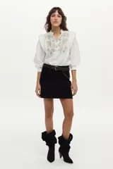 Blusa blanca de tela liviana con bordado detallado en el frente, cuello alto con volado y escote en V con botones. Tiene mangas largas abullonadas con puños amplios y detalles estructurados en los hombros.