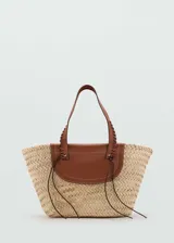 Bolso tipo cesta de fibras naturales con solapa y asas de cuero vacuno en color marrón. Presenta un diseño tejido a mano con detalles de costuras decorativas en las asas y tiras finas colgantes.