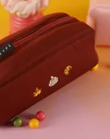 Neceser rectangular de tela color ladrillo con cierre superior y tres bordados pequeños: una hoja de arce, una taza de té y un croissant.