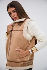 Chaleco de gamuza sintética color camel con interior y bordes de corderito sintético color crema. Presenta cierre frontal metálico, detalles de tiras con hebillas en el cuello y en la parte inferior, y costuras reforzadas con ribetes de cuero sintético.