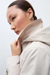 Trench coat de corte clásico en color beige, con cierre frontal de doble botonadura, solapas amplias y cinturón ajustable con hebilla. Incluye capucha desmontable y detalles de trabillas en los puños.