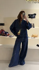Camisa de denim azul con cuello mao, diseño holgado, mangas largas y cierre frontal con botones metálicos. Cuenta con costuras decorativas pespunteadas en el delantero.