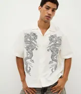 Camisa de sarga blanca de manga corta, con cuello inglés y cierre frontal de botones. Presenta un diseño boxy con estampa de dragones en el frente.