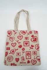 Bolso tote de tela con estampado de rayas verticales rosadas y blancas, laterales y asas en color beige. Presenta la palabra "BONJOUR" en verde en el centro y "INDIAN" en la parte inferior.