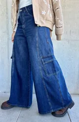 Pantalón tipo jean cargo de corte ancho y tiro alto, confeccionado en denim azul oscuro. Presenta bolsillos laterales tipo cargo con solapa y costuras reforzadas a lo largo de las piernas.