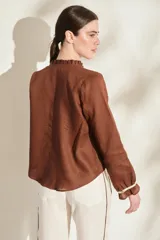 Blusa de lino color marrón con escote en V, cuello con volados y mangas largas con puños fruncidos que presentan detalles en color crudo, al igual que en los hombros. Cuenta con cierre frontal de botones.
