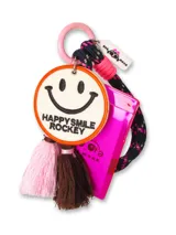 Llavero multifuncional con tarjetero de acrílico fucsia translúcido. Incluye un dije circular de tela con bordado de cara sonriente y la frase 'HAPPY SMILE ROCKEY', acompañado de borlas decorativas en tonos rosa y marrón. Se sujeta mediante una argolla metálica rosa y un cordón trenzado negro con detalles en rosa.
