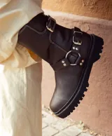 Bota biker de caña media confeccionada en cuero vacuno encerado. Presenta un diseño robusto con hebillas metálicas niqueladas en la caña y en la zona del tobillo, con una argolla decorativa. Cuenta con suela de goma negra con plataforma y cierre lateral interno.