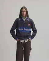 Sweater de punto Nude Project modelo Legend Football Knit Polo, con diseño a rayas en tonos azul y marrón. Cuenta con cuello polo con cierre frontal parcial, detalles bordados de una rosa y mariposas, y el nombre de la marca destacado en el pecho.