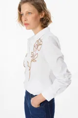 Camisa blanca de corte clásico con cuello camisero y cierre frontal de botones. Presenta un bordado lineal en color marrón oscuro en ambos lados del frente, representando motivos florales.