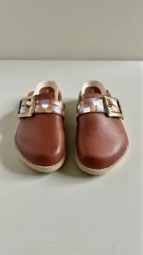 Sandalias de cuero color marrón de la marca Carolina de Cunto, modelo SAKI. Presentan una tira con hebilla metálica grande y detalles pintados a mano en tonos dorados, plateados y blancos. Cuentan con diseño destalonado y suela plana.