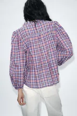 Blusa de manga larga con diseño a cuadros en tonos violeta, rosa y blanco. Presenta cuello redondo, cierre frontal con botones y mangas con frunces en los hombros.