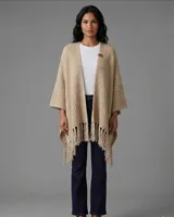 Ruana tejida en lana color beige, con diseño de punto texturizado, mangas amplias y terminación con flecos en el ruedo. Presenta una etiqueta de cuero decorativa en la parte superior.