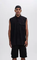 Camisa sin mangas de corte oversize, color negro, confeccionada en tejido twill. Presenta cuello clásico, cierre frontal oculto y un bolsillo tipo cargo en el pecho con detalle de remaches metálicos.