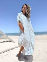 Vestido camisero midi de corte holgado, con estampado de rayas verticales celestes sobre fondo blanco. Presenta cuello camisero, cierre frontal con botones, mangas cortas con detalle de trabilla y bolsillos laterales.