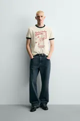 Camiseta de corte regular fit, color crema, con cuello redondo y mangas cortas. Presenta un estampado frontal con un personaje de dibujos animados jugando al baloncesto y texto "Hudson Valley Company Champion '87 NYC, USA". Los acabados del cuello y las mangas son de vivo negro a contraste.