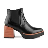 Bota tipo Chelsea de caña corta en color negro, confeccionada en material sintético con acabado brillante. Presenta una plataforma delantera negra con detalle de borde color madera y un taco cuadrado alto con textura símil madera.