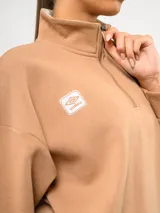 Buzo de felpa color tostado con cuello alto y medio cierre frontal. Presenta un diseño de corte holgado con hombros caídos, puños y ruedo acanalados, y el logo de la marca bordado en el pecho.
