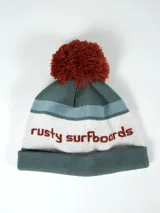 Gorro de lana Rusty Balve con pompón marrón, diseño a rayas en tonos verde, celeste y blanco, y texto bordado de la marca.
