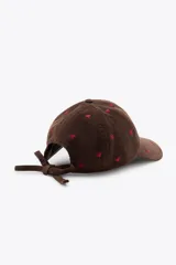 Gorra con visera de pana color marrón, con bordados de corazones rojos distribuidos por toda la superficie. Cuenta con cierre ajustable en la parte posterior.
