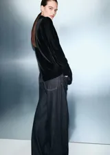 Blazer de terciopelo negro con cuello de solapa, cierre frontal cruzado con botones y mangas largas con puños abotonados. Presenta un diseño recto con bolsillos de solapa en la parte frontal y forro interior.