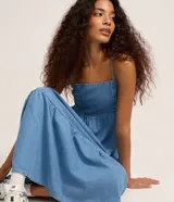 Vestido midi de jean azul con breteles finos, escote recto y falda con volados escalonados. Presenta un ajuste elástico en el busto y diseño holgado.