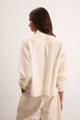 Camisa de algodón color crudo, con calce relajado, cuello clásico, botones delanteros, mangas largas con sutil volumen y ruedo asimétrico.