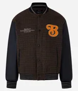 Campera bomber de estilo college con cuerpo a cuadros en tonos marrones y mangas largas de cuero sintético negro. Presenta cuello, puños y cintura acanalados a rayas, cierre abotonado, bolsillos laterales y aplique bordado de letra "B" en el pecho.
