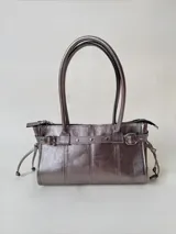 Cartera de cuero metalizado en tono plateado, de estructura rígida y tamaño medio. Presenta doble asa de hombro, cierre superior, y detalles decorativos que incluyen una tira horizontal con tachas y hebillas laterales, además de lazos ajustables en los costados.