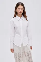 Camisa blanca de algodón, holgada, de manga larga con puños anchos, cuello camisero y cierre frontal oculto con un botón dorado visible en el cuello.