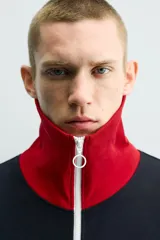 Chaqueta deportiva de corte regular, confeccionada en tejido piqué con interior forrado en red. Presenta un diseño color block en negro y rojo con detalles en blanco, cuello subido, cierre frontal con cremallera y bolsillos laterales con cierre.