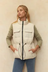 Chaleco puffer color crema, con cuello alto, cierre frontal y cuatro bolsillos con solapa. Cuenta con cintura ajustable con cordón.