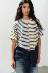 Camiseta oversize con diseño bicolor a rayas horizontales, combinando un lado en tonos rosado y verde oscuro, y el otro en beige y marrón. Cuenta con cuello redondo, mangas cortas amplias y texto bordado en azul en el pecho.