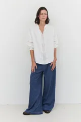 Camisa blanca de corte holgado con cuello mao y escote en V con cordones terminados en borlas. Presenta detalles de bordados calados verticales en el frente y en las mangas, con mangas largas abullonadas y puños elásticos.