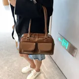 Cartera de cuero sintético color marrón, de diseño rectangular con doble asa superior para hombro, cierre principal con cremallera y dos bolsillos frontales externos con solapa.