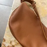 Bandolera de cuero graneado color marrón, con forma de media luna y correa ajustable.
