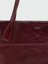 Bolso shoulder bag de Balmain en color bordó, confeccionado en cuero, con doble asa superior y correa ajustable para el hombro. Presenta costuras decorativas y el logo de la marca estampado en tono dorado en la parte frontal.