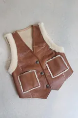 Chaleco de cuero genuino color tostado con interior y detalles en piel sintética color crema. Presenta cierre frontal con botones, dos bolsillos delanteros y diseño con costuras visibles.
