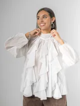 Camisa blanca de algodón italiano con cuello alto ajustable mediante lazo trasero, mangas abullonadas y un gran shabot frontal con volados en cascada.