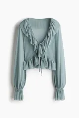 Blusa corta confeccionada en malla transparente de color verde, con escote en V pronunciado, tiras finas para anudar en el frente y volados escalonados en el escote y el bajo. Presenta mangas largas con puños elásticos y acabados acampanados.