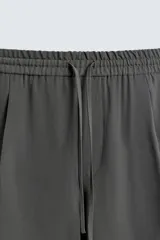 Pantalón de corte relajado, color gris oscuro, con cintura elástica ajustable mediante cordón y pliegues frontales. Presenta bolsillos laterales y bolsillos traseros de parche con pespuntes contrastantes.